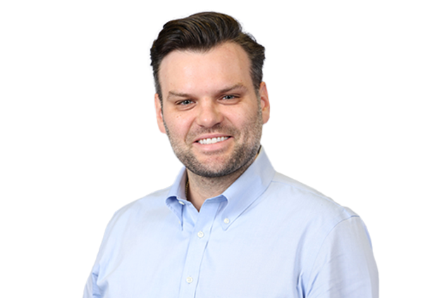 A man with dark hair and a beard is smiling while wearing a light blue button-up shirt against a white background.
