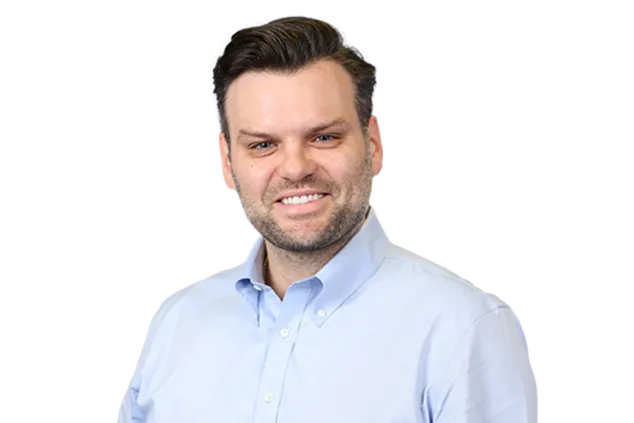 A man with dark hair and a beard is smiling while wearing a light blue button-up shirt against a white background.