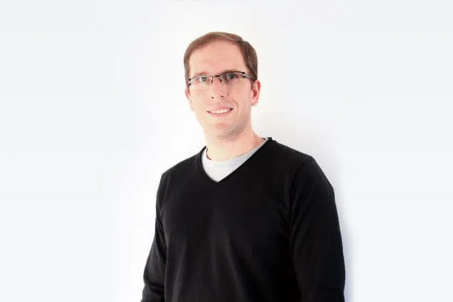 A man wearing glasses and a black sweater stands against a plain white background, looking towards the camera and smiling.