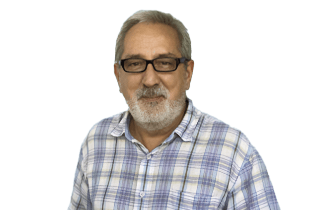 An older man with short gray hair, glasses, and a white beard, wearing a blue and white plaid shirt, stands against a plain white background.
