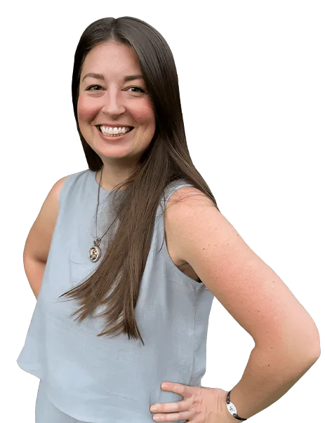 A woman with long brown hair, wearing a sleeveless gray top, stands smiling with her hands on her hips. She has a necklace and a watch.