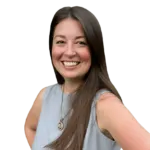 A woman with long brown hair is smiling and wearing a sleeveless grey top and a necklace. She is posing with one hand on her hip against a plain white background.