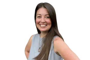A woman with long brown hair is smiling and wearing a sleeveless grey top and a necklace. She is posing with one hand on her hip against a plain white background.