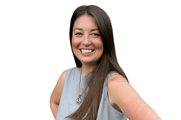A woman with long brown hair is smiling and wearing a sleeveless grey top and a necklace. She is posing with one hand on her hip against a plain white background.