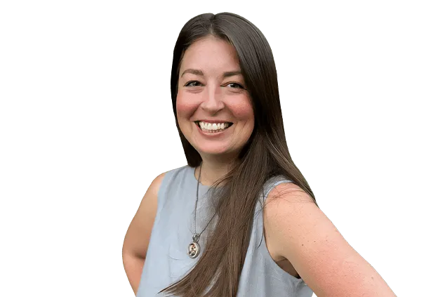 A woman with long brown hair is smiling and wearing a sleeveless grey top and a necklace. She is posing with one hand on her hip against a plain white background.