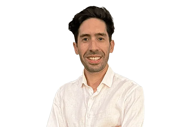 A man with dark hair and a beard, wearing a white button-up shirt, stands against a plain background and smiles at the camera.