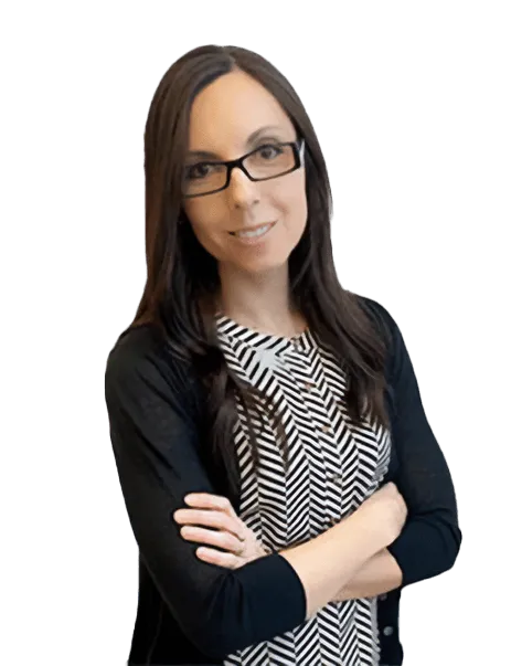 A woman with long dark hair and glasses stands with arms crossed, wearing a patterned dress and a black cardigan.