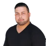 A man with short dark hair and a beard, wearing a black V-neck shirt, looks directly at the camera against a white background.