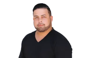 A man with short dark hair and a beard, wearing a black V-neck shirt, looks directly at the camera against a white background.