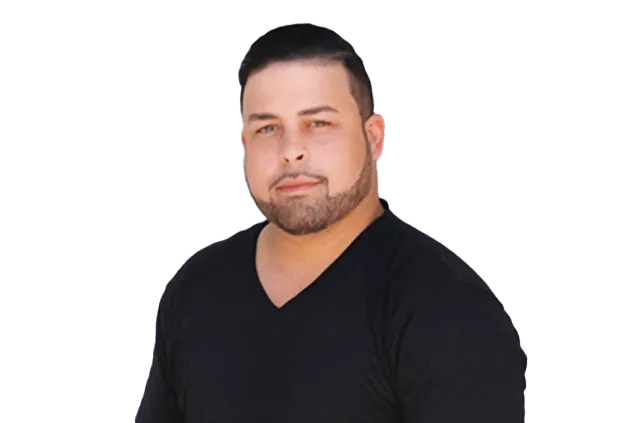 A man with short dark hair and a beard, wearing a black V-neck shirt, looks directly at the camera against a white background.