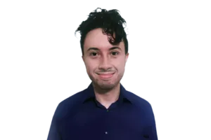A person with dark curly hair wearing a blue shirt, smiling at the camera against a plain, dark background.