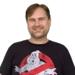A man wearing a black "Ghostbusters" T-shirt smiles while standing against a white background.