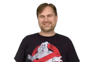 A man wearing a black "Ghostbusters" T-shirt smiles while standing against a white background.