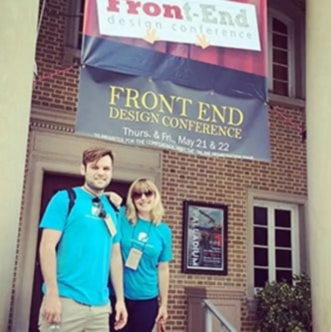Two people in blue shirts stand in front of a building under a banner that reads "Front-End Design Conference." They appear to be attending the event.