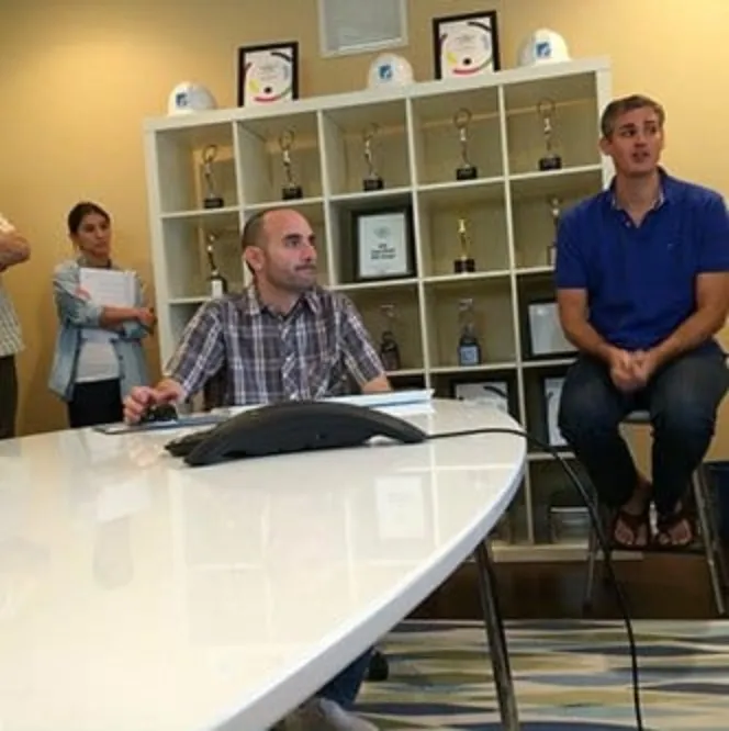 Four people in a room with a large white table and shelves displaying awards. Two seated men are in the foreground; a man and a woman stand in the background near the shelves.
