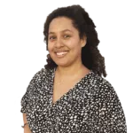 A person with curly hair, wearing a black and white patterned blouse, smiles at the camera.