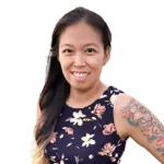 Woman with long dark hair and floral dress smiling, displaying a colorful sleeve tattoo on her left arm. Background is plain white.