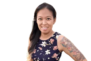 Woman with long dark hair and floral dress smiling, displaying a colorful sleeve tattoo on her left arm. Background is plain white.