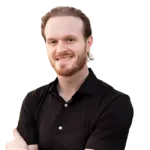 A man with light brown hair and a beard, wearing a black shirt, stands with arms crossed, smiling at the camera against a plain background.