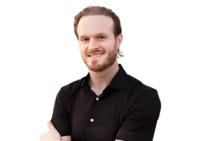 A man with light brown hair and a beard, wearing a black shirt, stands with arms crossed, smiling at the camera against a plain background.
