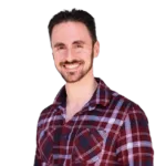 A man with dark hair and a beard smiles at the camera. He is wearing a red plaid shirt.