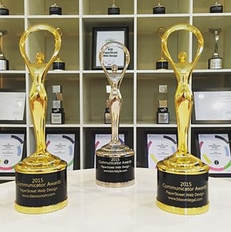 Three 2015 Communicator Awards trophies are displayed on a shelf, with other award plaques and certificates visible in the background.