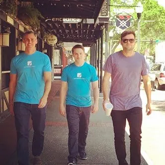 Three men walk down a sunlit sidewalk, two wearing matching blue t-shirts and one in a purple t-shirt, with storefronts and trees lining the street in the background.