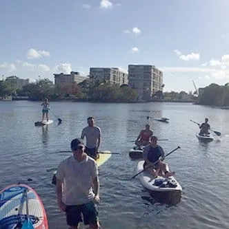 A group of people is paddleboarding and kayaking on a calm water body with buildings and trees visible in the background.