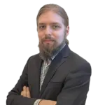 A bearded person with long hair is wearing a dark suit and has their arms crossed, looking at the camera against a white background.