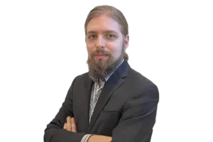 A bearded person with long hair is wearing a dark suit and has their arms crossed, looking at the camera against a white background.