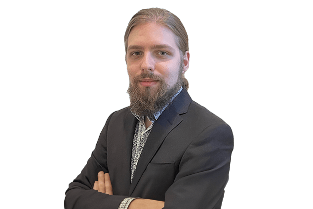 A bearded person with long hair is wearing a dark suit and has their arms crossed, looking at the camera against a white background.