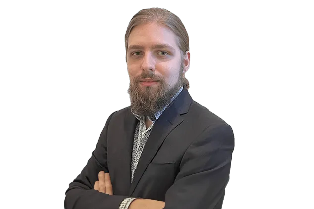 A bearded person with long hair is wearing a dark suit and has their arms crossed, looking at the camera against a white background.