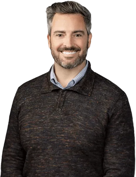 A man with short, graying hair and a beard is smiling. He is wearing a dark, textured sweater over a light-colored collared shirt.