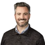 A man with gray hair and a beard is smiling. He is wearing a dark sweater over a collared shirt. The background is transparent.