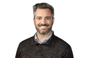 A man with gray hair and a beard is smiling. He is wearing a dark sweater over a collared shirt. The background is transparent.