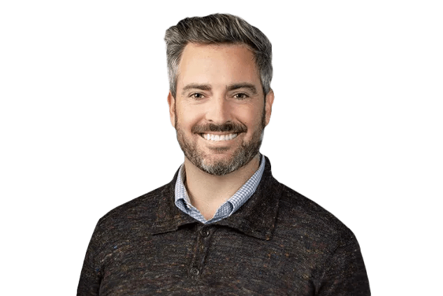 A man with gray hair and a beard is smiling. He is wearing a dark sweater over a collared shirt. The background is transparent.
