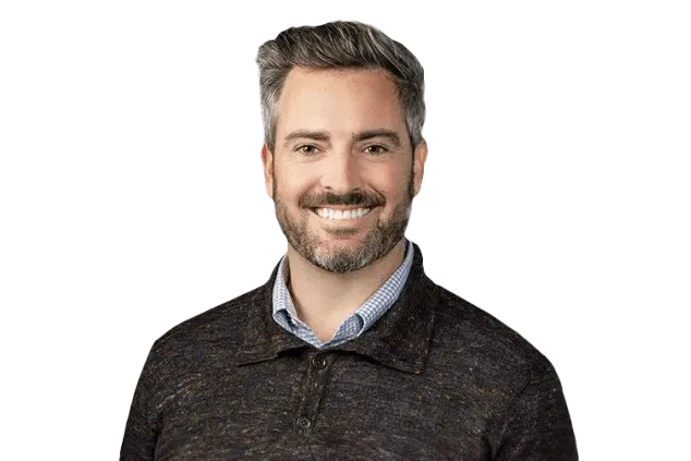 A man with gray hair and a beard is smiling. He is wearing a dark sweater over a collared shirt. The background is transparent.