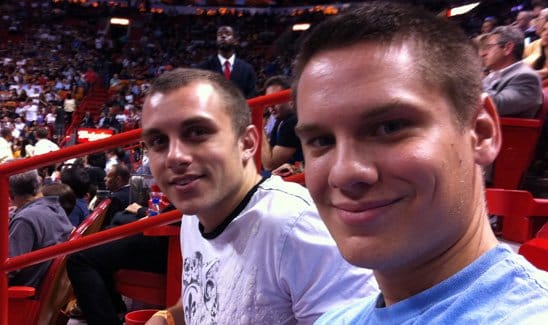 Two young men are sitting in the stands at a sports event, with the arena crowd visible in the background. One wears a light-colored t-shirt, and the other wears a blue t-shirt.