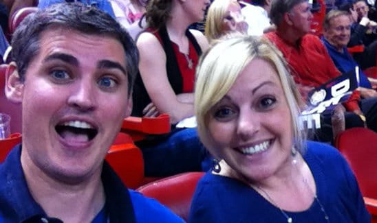 A man and a woman are smiling at the camera while seated in a crowded place, with other people visible in the background.