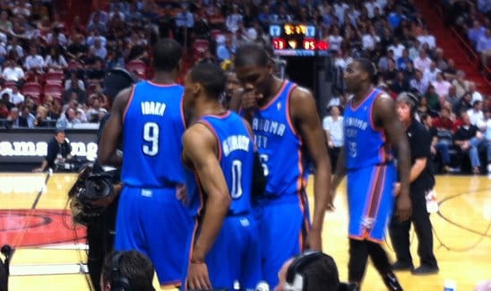 A group of basketball players in blue "Oklahoma City" uniforms are gathered on the court during a game, with a crowd visible in the background.