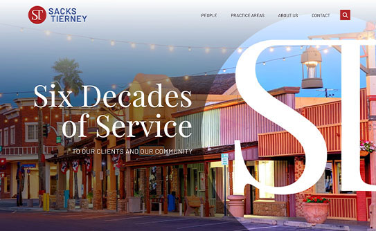 Website screenshot of Sacks Tierney law firm with the text "Six Decades of Service to Our Clients and Our Community" overlaid on a background image of a row of shops and streetlights.