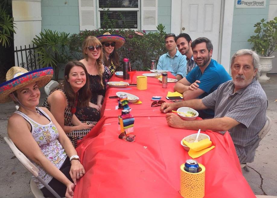 A group of eight people sitting around a table outdoors, sharing a meal. The table has a red tablecloth and some are wearing sombreros.
