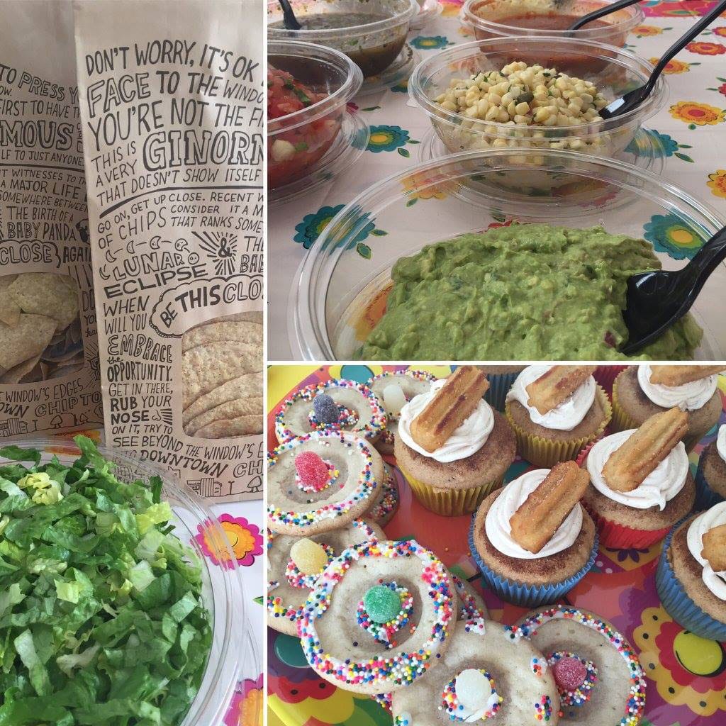 A table with food items including bowls of guacamole, salsa, corn, and lettuce, bags of tortillas, and a variety of cupcakes and cookies decorated with colorful frosting and toppings.