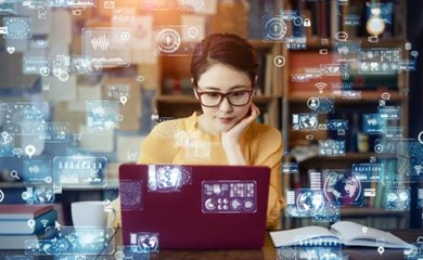 A woman wearing glasses sits at a desk with a laptop, surrounded by holographic data and technology icons, implying digital interaction or data analysis. She appears focused and engaged.