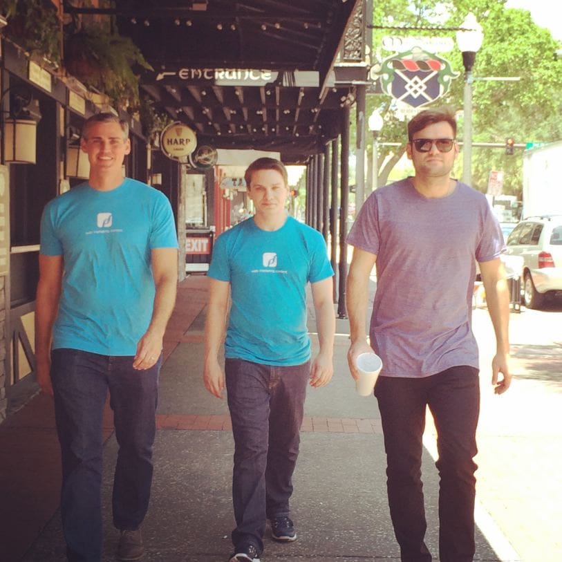 Three men walking down a sidewalk in an urban area. Two of them are wearing matching blue t-shirts, and one is wearing sunglasses and holding a white cup. Greenery and storefronts are in the background.