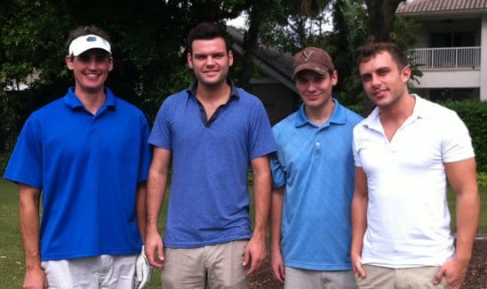 Four men are standing outside, smiling at the camera. They are casually dressed in polo shirts and shorts, with trees and a building in the background.