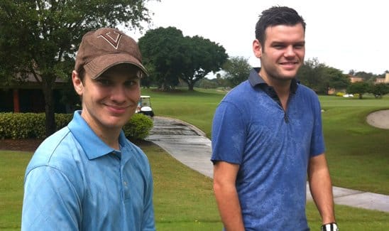 Two men standing on a golf course, smiling at the camera. One is wearing a baseball cap and both are in casual attire suitable for golfing. The background shows greenery and a golf cart path.