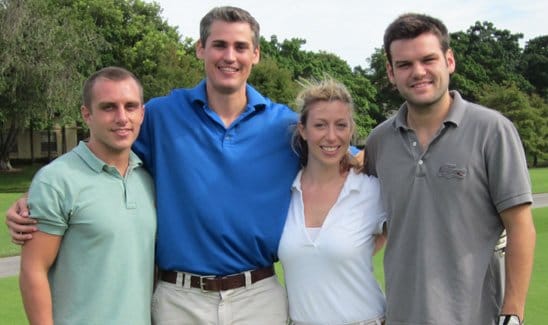 Four people are standing together outdoors on a grassy area, smiling at the camera.