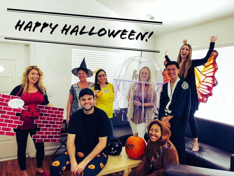 Group of eight people wearing various Halloween costumes, posing indoors with a "Happy Halloween!" banner above, and festive decorations around them.