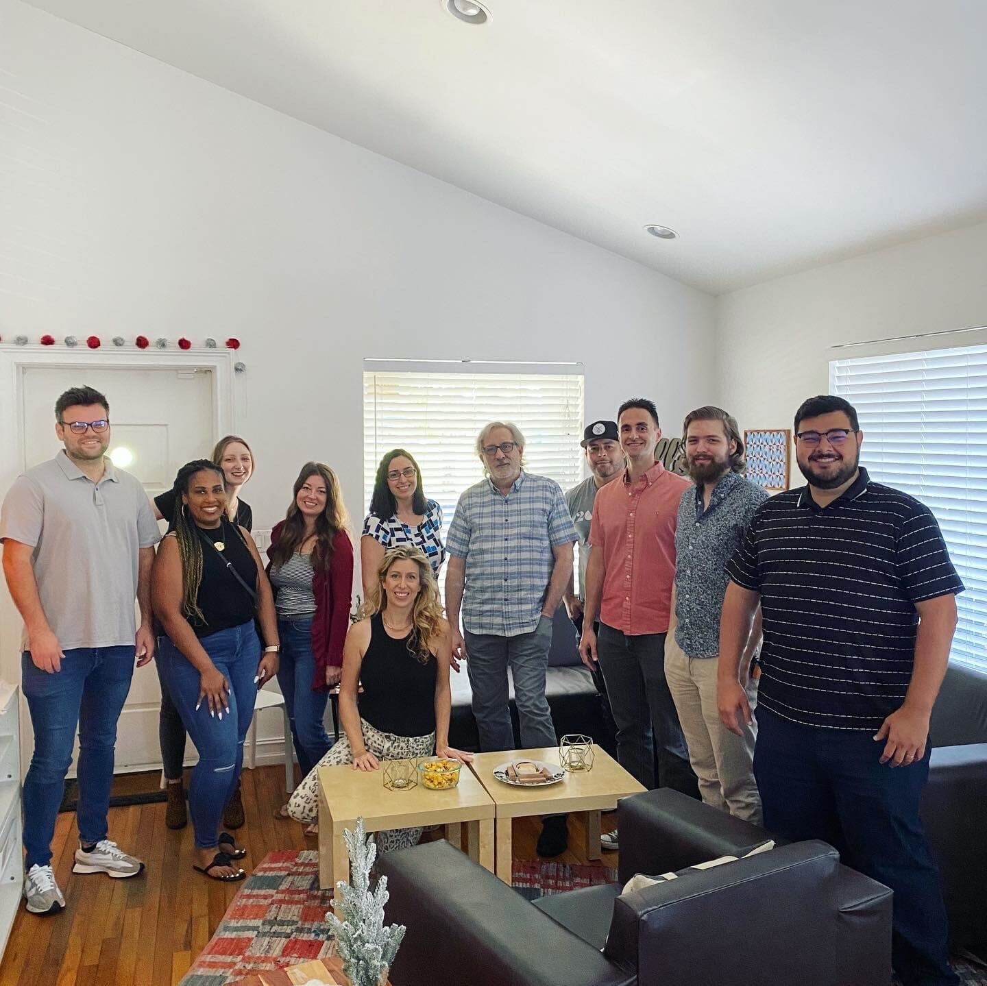 A group of eleven people posing in a brightly lit living room. Two people are seated on couches, and nine are standing behind them. A coffee table is in the center with snacks.